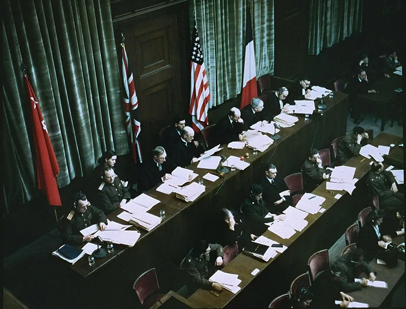 Three rows of judges sit at long tables in front of Soviet, British, American, and French flags, examining numerous sheets of paper.