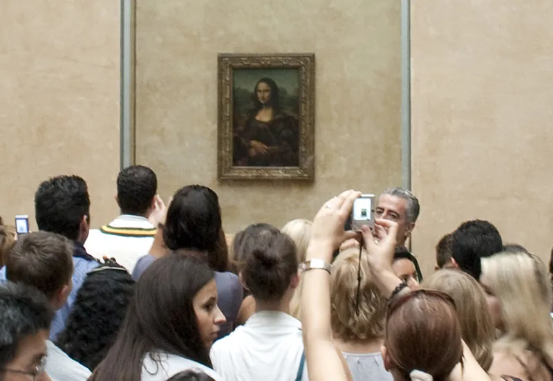 A crowd of museum goers viewing the Mona Lisa. One takes a photograph with a cell phone.