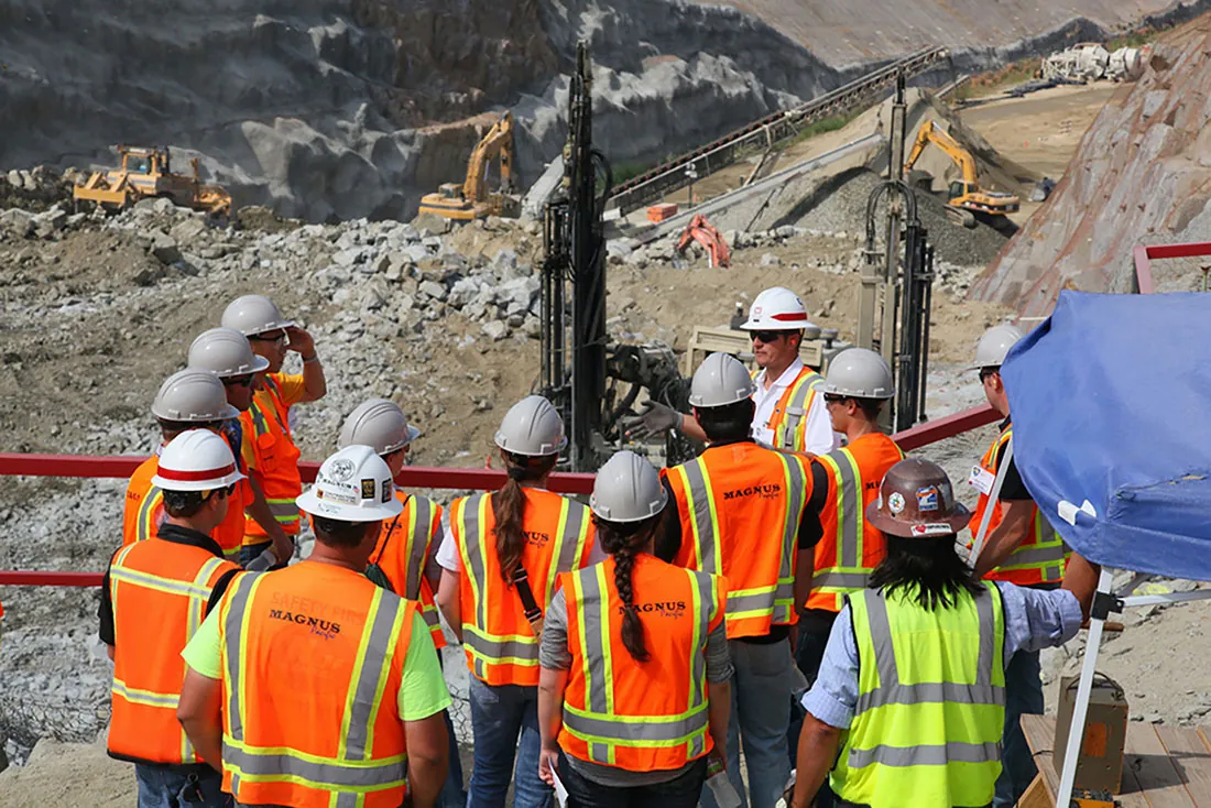 Students wearing bright orange and yellow construction vests are shown standing around an outdoor job site.