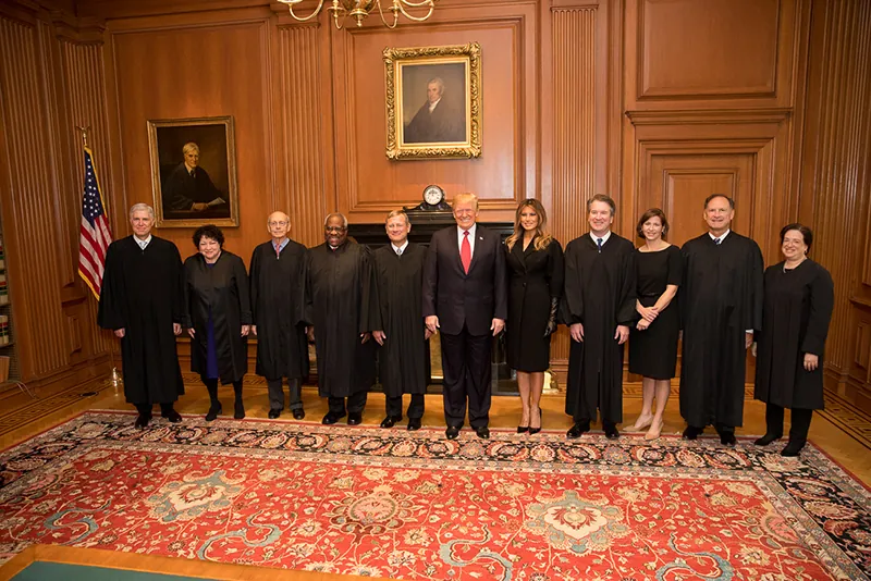 Members of the US Supreme court stand for a portrait with Donald Trump and Melania Trump in a wood-paneled room in front of a fireplace.