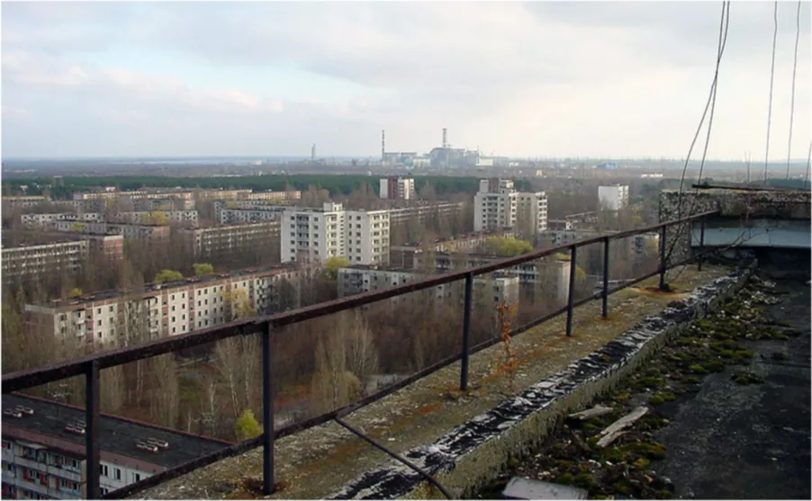 A picture shows the view from the top of a building. There is a metal fence around the edges of the building in the concrete roof and wires going up from the fence on the right side. The floor of the roof looks dark and littered. The view shows tall and long white buildings and green trees in front of a blue sky. In the background there is a large group of buildings with tall pipes.