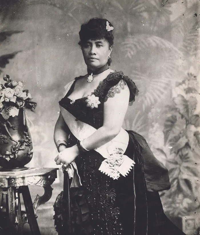 A woman is shown in a black and white photograph. She is wearing a low cut beaded dark dress with lacy cap sleeves. A brooch hangs from a beaded ribbon around her neck, her hair is up, adorned with a butterfly pin and she wears earrings, rings, and bracelets. A white sash runs from her right shoulder and ends at her left waist with a circular ornament. She holds white gloves in her hands and leans on a cane. In the left of the picture is a table with a round, carved vase filled with flowers. The wall behind her is decorated with leaves and plants.