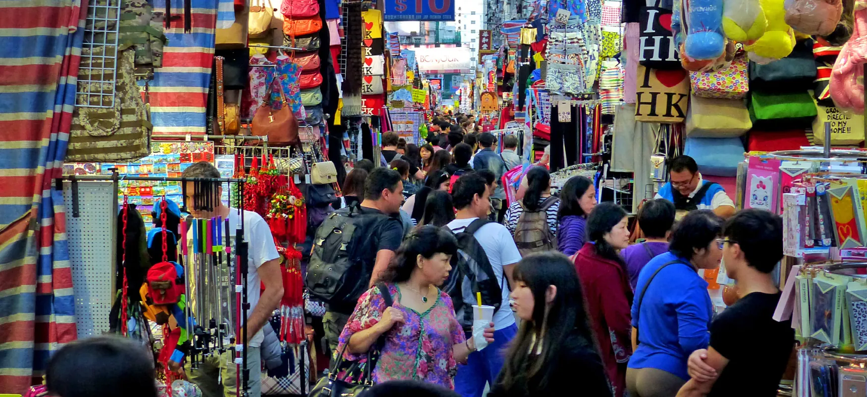 A large crowd of people walk through a street market. Bags, jewelry, and other items available for sale are visible.