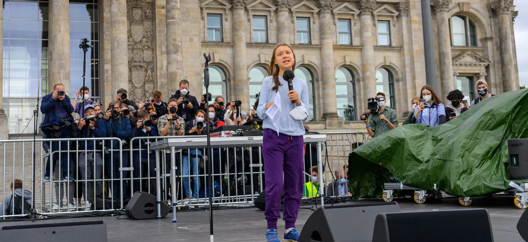 Greta Thunberg addressing a crowd in front of a governmental building with reporters and photographers filming and photographing from behind the stage.