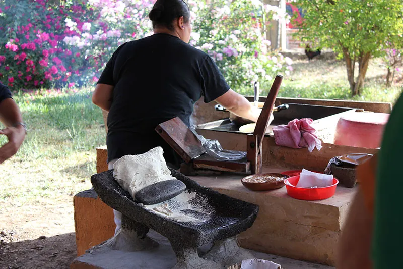 A woman doing work with her back to the camera, with a stone grinder and grindstone visible in the foreground.