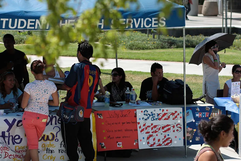 People sitting at tables during a college club showcase fair, while prospective members speak to them and look at materials. The tables are decorated with signs for clubs including the Student Veteran’s Club.