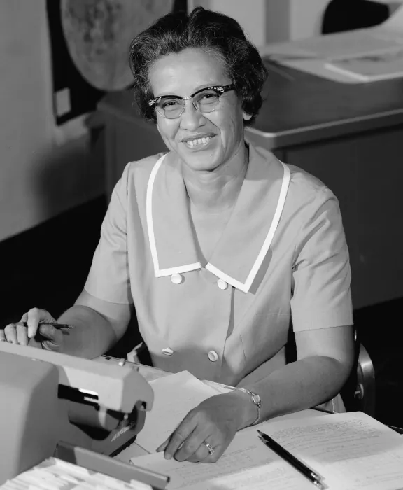 A photo of mathematician Katherine Johnson seated at a desk with what appears to be a manual calculation machine and a number of papers with tables on them. 
