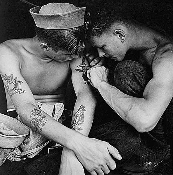 Black and white image of two young sailors, one tattooing the arm of the other.