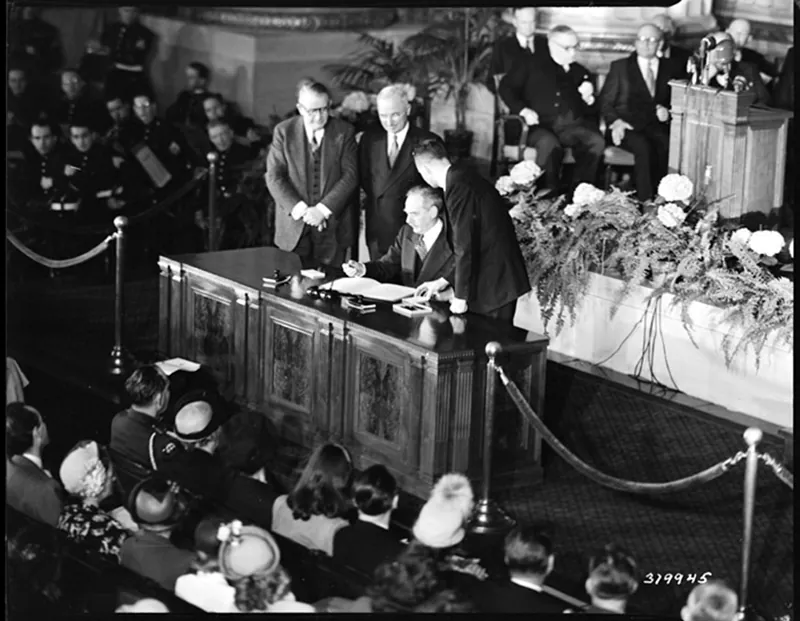 President Harry Truman and Vice President Alben Barkley stand together on a dais in front of a table festooned with plants and flowers, looking on while U S Secretary of State Dean Acheson signs a document. A crowd sits in rows around them.