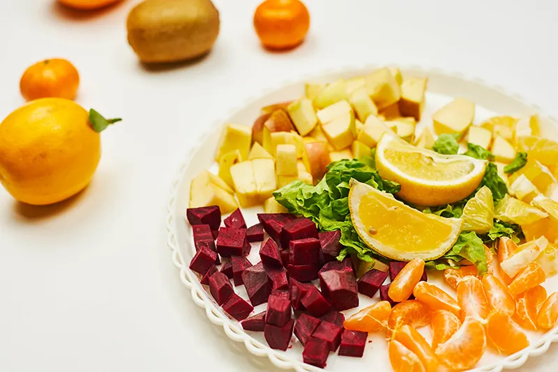 A plate of cut up beets, apples, and oranges. On the table behind the plate are several whole fruits.
