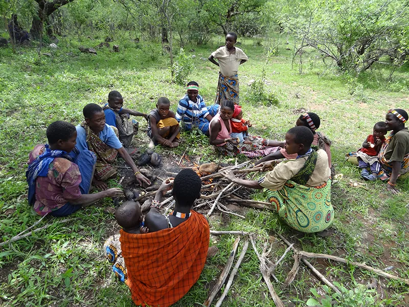 A group of 10 woman, seveal with babies strapped to them, sitting around a fire cooking food.