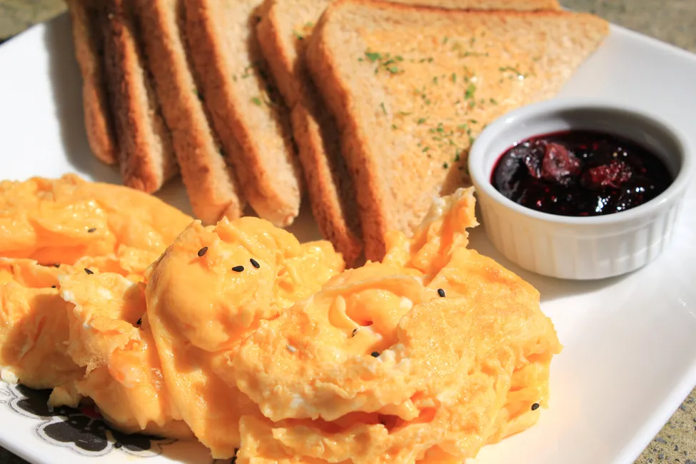 Se muestra una foto de un plato con un gran montón de huevos en primer plano y seis rebanadas de pan tostado en el fondo. Hay un pequeño plato de mermelada roja cerca del pan tostado en el plato.
