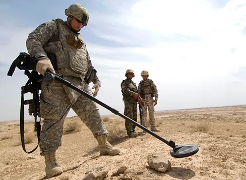 Zdjęcie przedstawia żołnierza armii Stanów Zjednoczonych, używającego wykrywacza metali do poszukiwania materiałów wybuchowych i broni w pustynnym terenie Iraku
