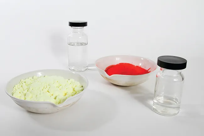 This photo shows two vials filled with a colorless liquid. It also shows two bowls: one filled with an off-white powder and one filled with a bright red powder.