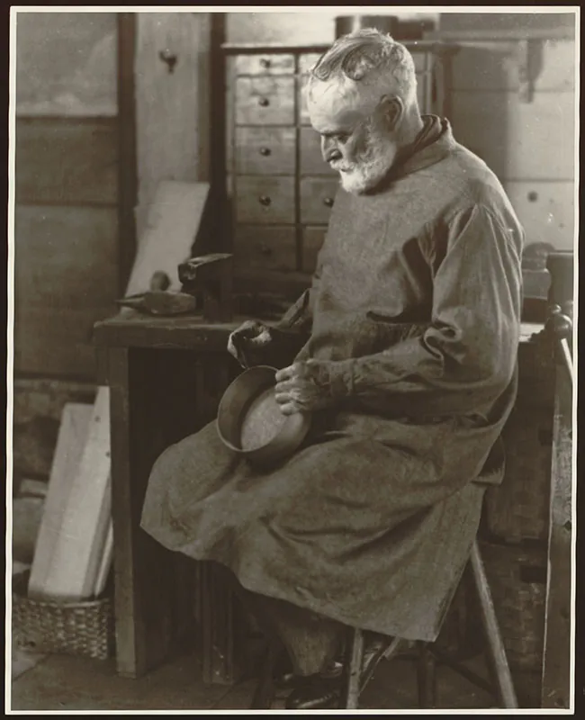 A man sits on a bench holding an oval box in his hands. He wears a knee-length smock. Wood working tools are visible on the table behind him.