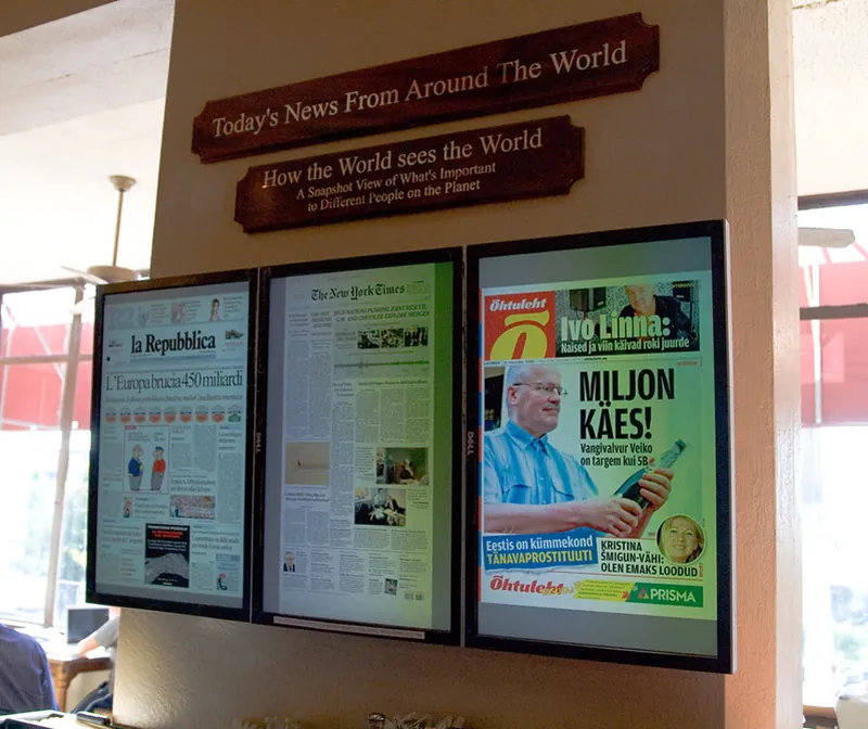 Three rectangular screens displayed side-by-side on a wall show rotating front pages of newspapers from around the world. Signs on the wall above the screens read: “Today’s News From Around the World” and “How the World sees the World: A Snapshot View of What’s Important to Different People on the Planet.”