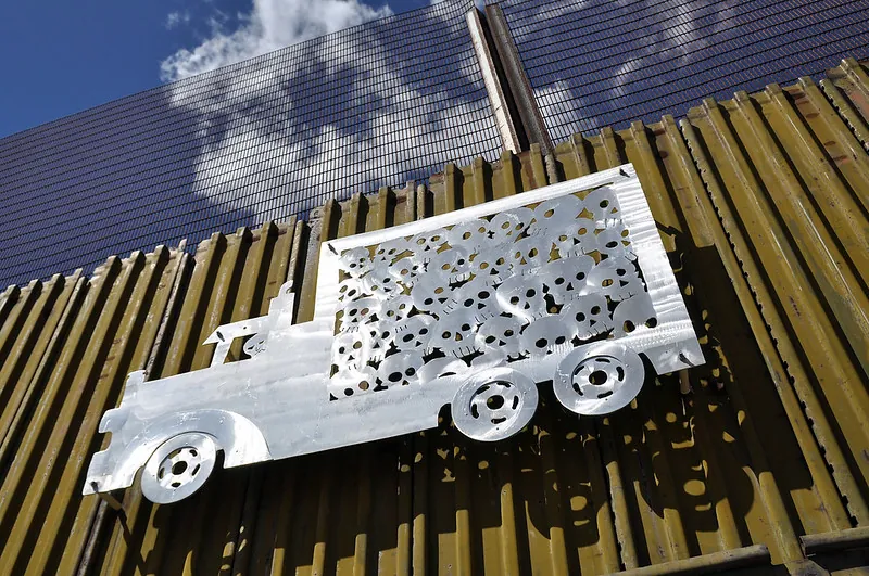 Color photograph of a two-dimensional box truck sculpture mounted to a rusty metal wall. The art seems to be cut out of a single piece of aluminum and shines brightly against the dingy background. The back of the truck is filled with cut-out images of a human skulls and another skull peers out from the driver’s window.