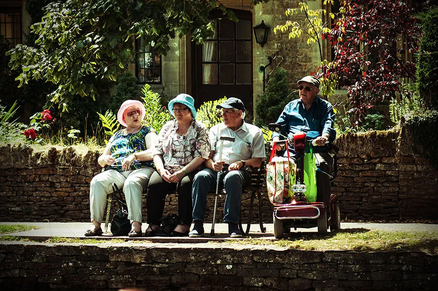 Three elderly people sit on a bench, one holding a cane. Next to them a fourth person sits on a scooter.