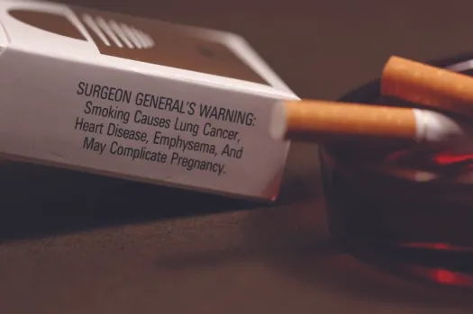 A photo of a cigarette box and two cigarettes. The cigarettes are resting in an ashtray. Text on the cigarette box reads “Surgeon General’s Warning: Smoking causes lung cancer, heart disease, emphysema, and may complicate pregnancy”.
