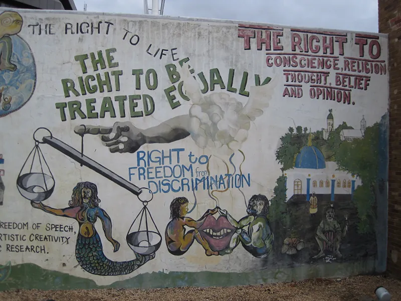 A mural is painted on an outdoor wall. The mural shows a mermaid holding the scales of justice, two people sitting on either side of a pair of lips, and a building surrounded by trees. Surrounding these images are the words “The right to life;” “The right to be treated equally;” “Freedom of speech, artistic creativity, and research;” “Right to freedom from discrimination;” and “The right to conscience, religion, thought, belief, and opinion.”
