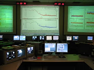 The image shows the LIGO Hanford control room. There are desktop computers and large screens showing a variety of graphs, likely showing spectra collected from stars.
