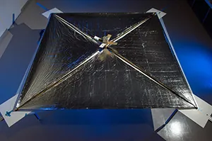 An image of the NanoSail is shown. The sail is shown fully unfurled, with the satellite in the center. The width of the satellite is approximately one-eighth the width of the sail.