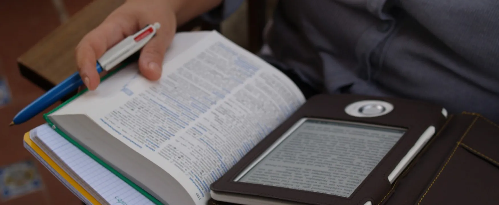 A close-up of a person reading with a tablet e-book device on top of a printed book. The printed book has underlines and notes in the margin in blue ink. Underneath the printed book is a notebook.