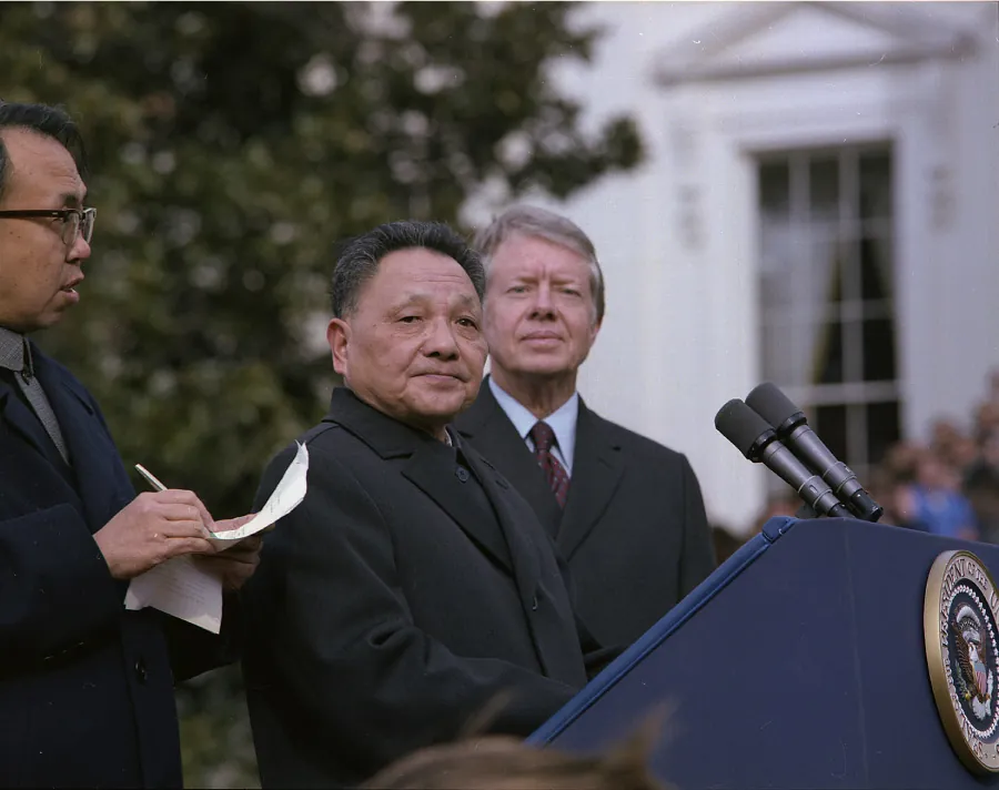 A photograph shows three men standing outside behind a podium with microphones. The man on the far left holds a paper and pen in his hand and looks to the man in the center. The man in the center stands behind the podium and looks at the camera. Behind him stands another man who also looks forward.