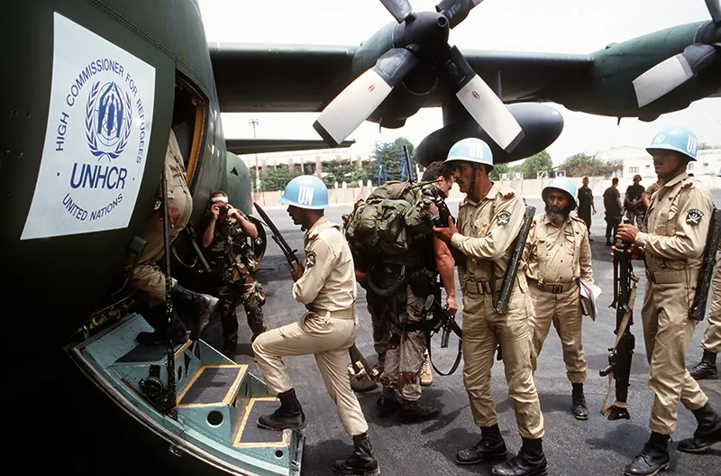 U N troops in blue helmets and tan uniforms and carrying weapons board a UN Refugee Agency aircraft.