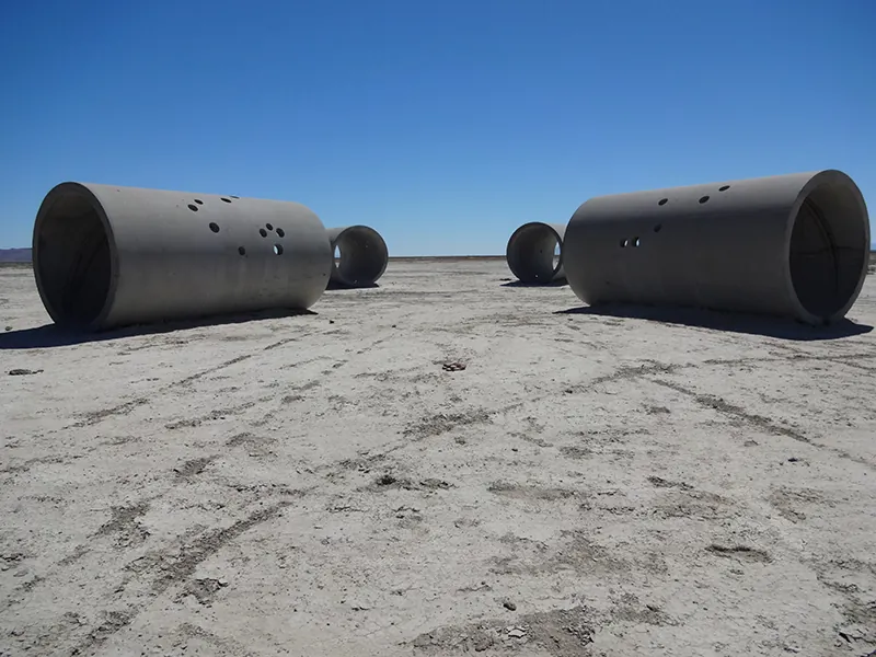 Photograph of four huge concrete cylinders positioned in an x-formation in a barren dessert setting. In addition to the openings at the front and the back, the cylinders have small round holes scattered along their tops and sides.