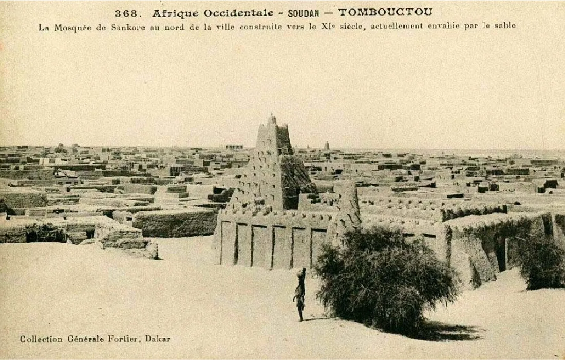 The postcard shows a brown, sandy mosque with tall walls and a seven-story building in the middle showing above the walls. No openings are visible in the walls. In the background a city with low buildings is shown. A person is visible in the foreground standing next to one of two bushes.