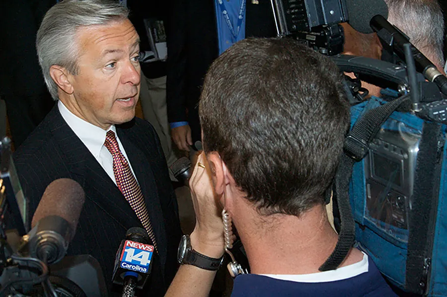 Wells Fargo CEO John Stumpf speaks to reporters who are crowding around him holding microphones and cameras.
