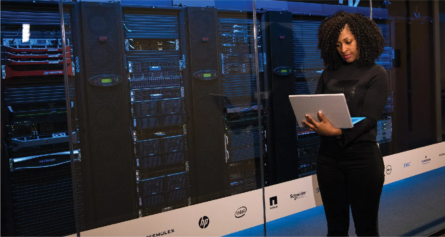 A person holds a laptop while standing in front of several racks of servers.