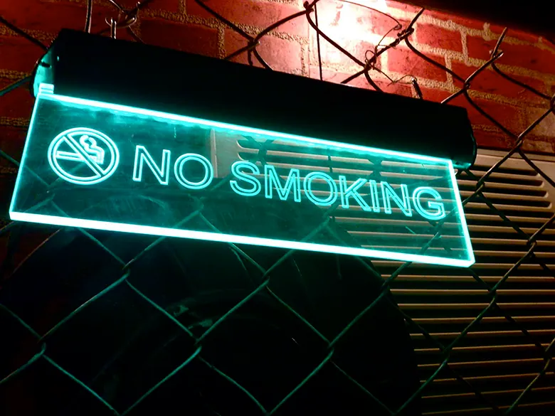 A photo shows an illuminated “No Smoking” sign mounted on a fence.