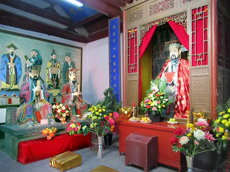 Interior of temple. One wall is taken up by a large status of Confucius, set into a recess lined with red curtains. Other statues appear on one side, as well as a large painting of several men. Large vases of flowers stand on the floor and on platforms. A box with a slot for offerings is in front of the statue of Confucius. Cloth covered rectangles to kneel upon are on the floor.