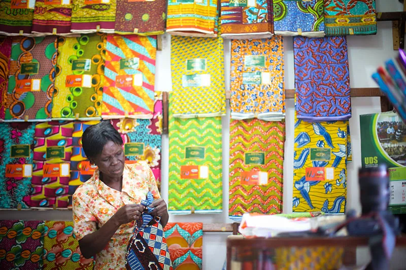 A color photograph of a woman standing in front of a wall displaying lengths of colorful fabric decorated with large, vibrant designs. The woman holds some fabric, which she appears to be hand-sewing.
