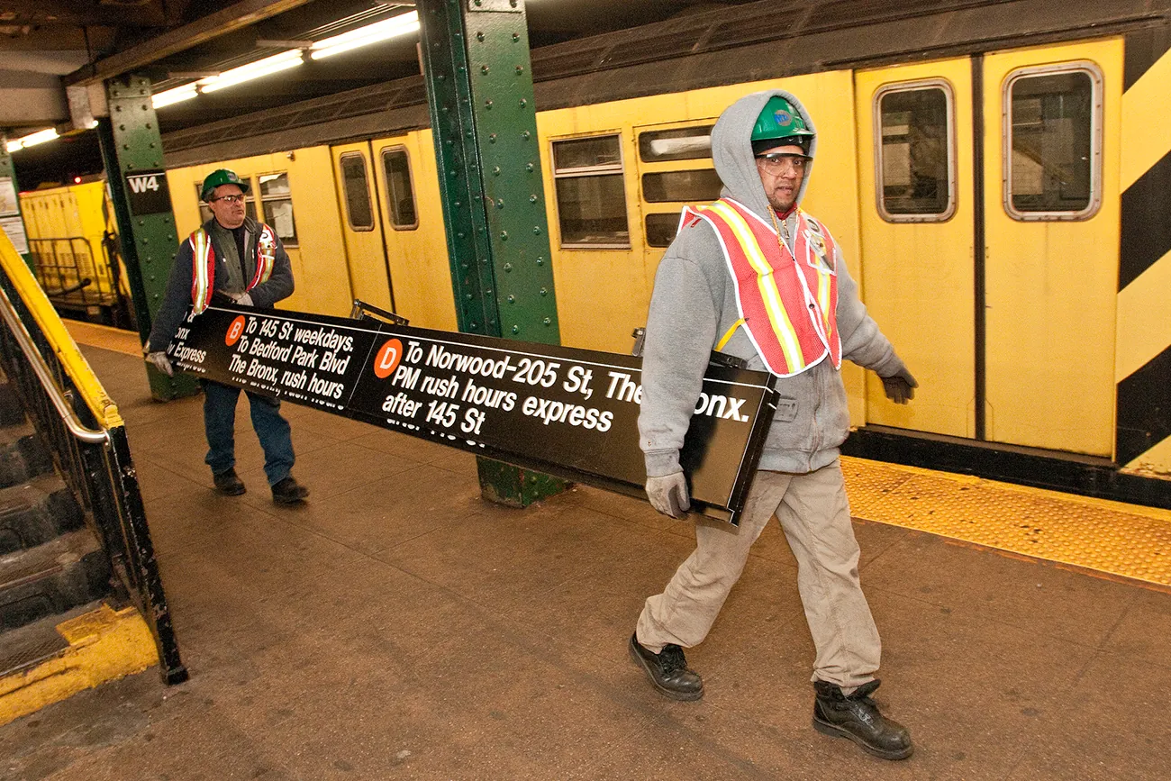 A photo shows two metro workers at work. They carry a destination sign inside a subway station.