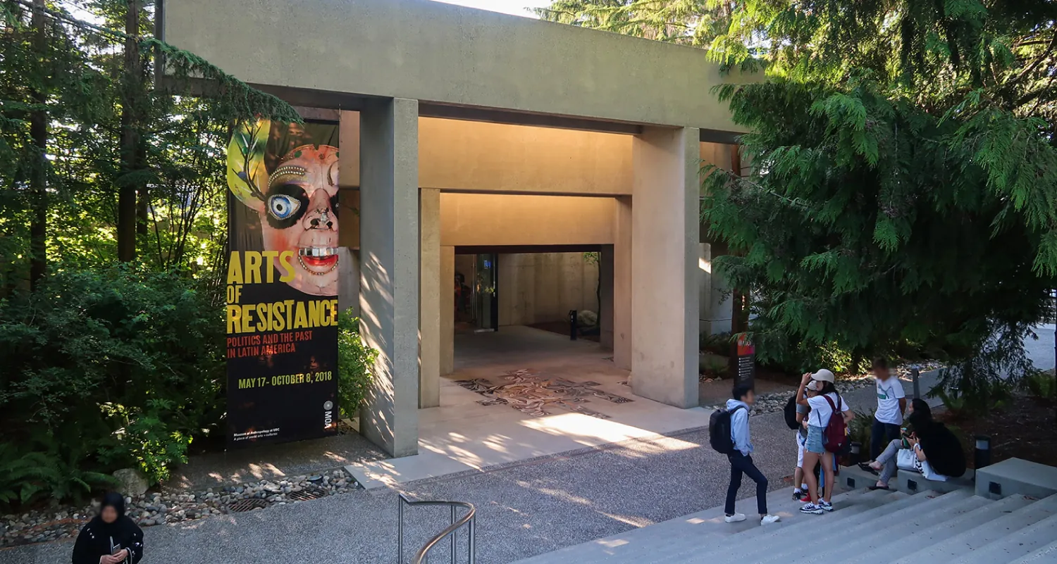 The front entrance to the Museum of Anthropology in British Columbia. The opening is a large rectangle outlined by rectangular cement columns of graduating sizes leading to the door. Modern in design. Sign on the left advertises an exhibit titled “Arts of Resistance”. Several people are gathered on the stairs in front of the building.