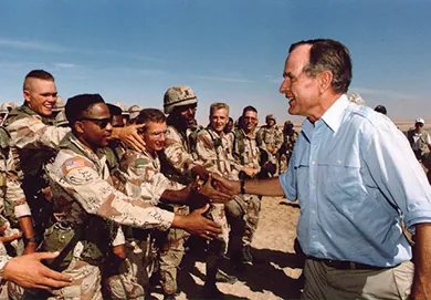 A photograph shows George H. W. Bush greeting and shaking hands with U.S. troops stationed in Saudi Arabia.