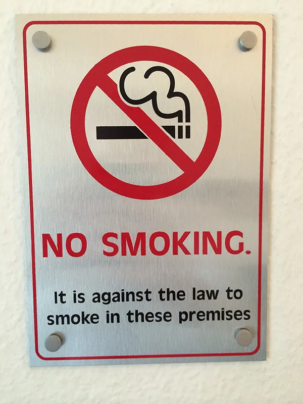 A metal sign shows a cigarette burning with a cross-out of the cigarette in red. Underneath the image are the words “No Smoking” and “It is against the law to smoke in these premises.”