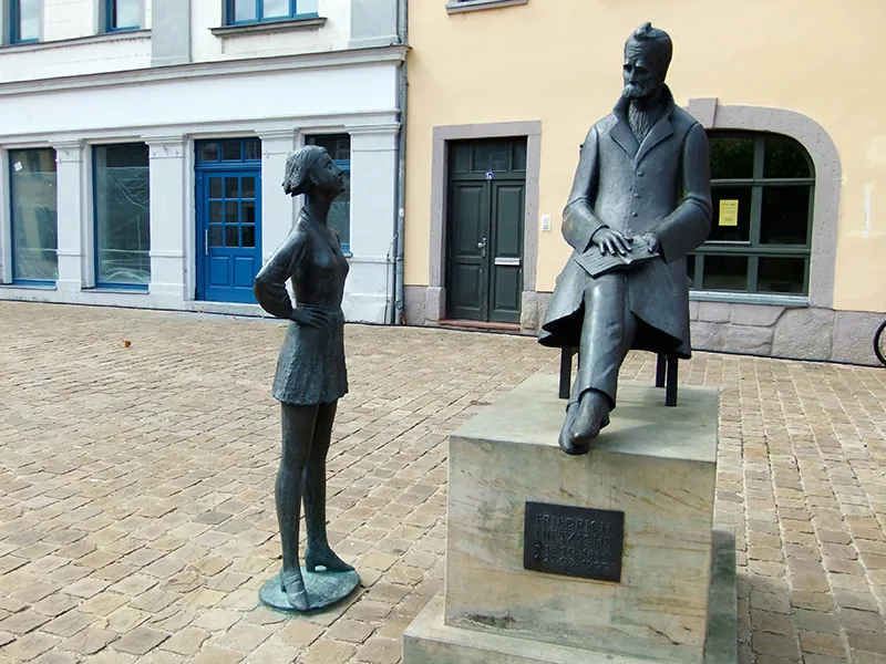 Public art consisting of two figures: a seated man with a book on his lap and a young woman in contemporary dress standing with her hands on her hips. The seated man is raised on a pedestal. The young woman is on the ground. The two figures look at one another.