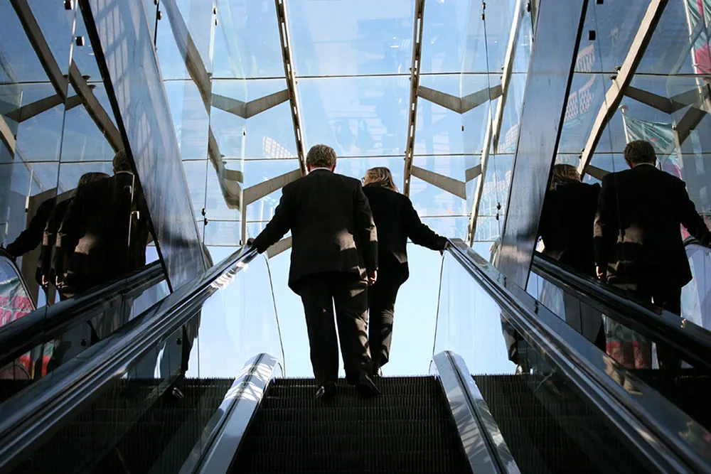 A man and a woman, both wearing business suits, are shown from behind at the top of an escalator