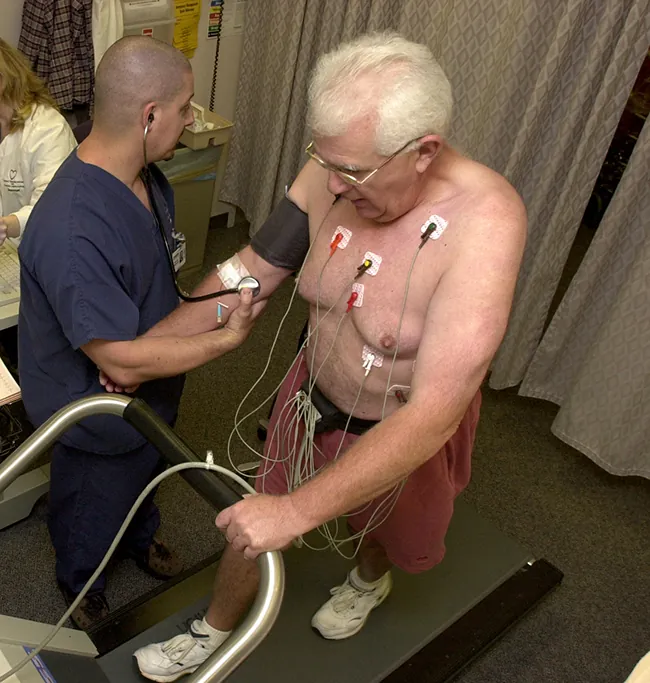 Se muestra una foto de dos hombres: uno caminando en una cinta de correr con varios cables conectados a su torso, y el otro recopilando los datos de la presión arterial del primero.