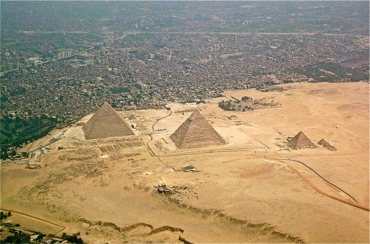 An aerial picture of three triangular sand-colored brick structures is shown on a sandy landscape with a city shown in the top of the picture. The two left structures are large and the third one on the right is smaller. Next to the smaller one are two small triangular structures with a third one shown in ruins. The sandy land surrounding the structures shows a road winding from the city to the bottom of the picture.