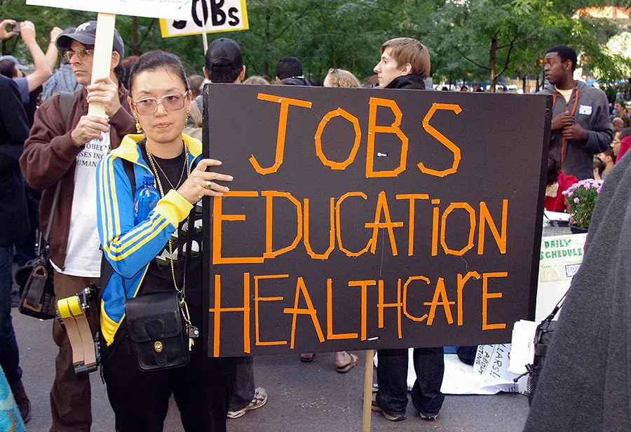 A person stands at a protest holding a large sign. The sign reads “Jobs, Education, Healthcare.”