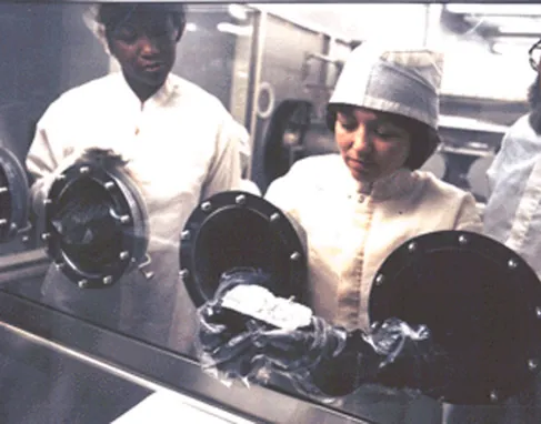 Photograph of Technicians Examining a Lunar Sample. Two scientists look at a Moon rock through the glass of a scientific “glove box”.