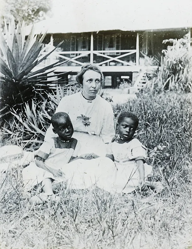 Black and white photograph of a White woman sitting in the grass in front of a house, with two young Black children sitting on either side. All three wear solemn expressions. A simple wood frame building is visible in the background.