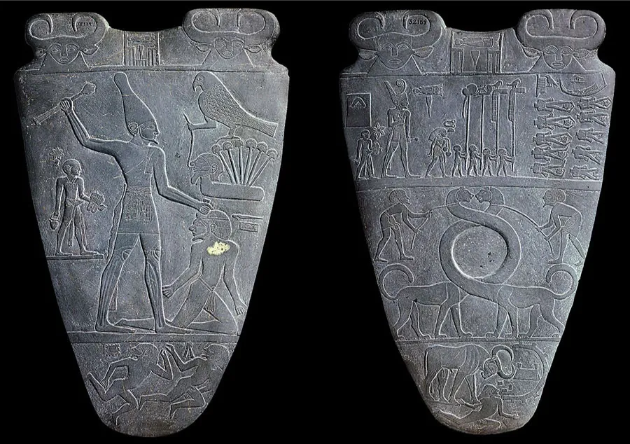 A picture of two arrow-shaped gray stones is shown on a black background. Both have rounded points along the bottom and along the longer top there are carving of bulls on either end, with their curly horns creating the shapes at the top of the stone. The stone on the left shows three rows of images. The top row shows two bulls with curly horns and etchings in between them. The second row shows a very tall person with a cloth at their waist, a tall, rounded hat on their head, a thin beard, bare-chested, and barefoot. He holds a long stick in his right hand and his left hand rests on the head of a man in a loincloth and headdress kneeling on the ground in front of him. Above the kneeling man is a bird standing on one leg on top of six tall mushroom-like projections coming out of a platform with a head at the left. His other foot is holding a stick that is attached to the nose of the head on the platform. To the left of the large man is another man standing in a loincloth holding objects with a sword at his side. The third row at the bottom shows two figures in minimal loincloths and headdresses running to the right and looking to the left. The stone on the right shows four rows of images. The top row shows two bulls with curly horns and etchings in between them. The second row shows four small people in the middle walking toward the right, holding tall staffs with various animals and items at the tops. To the left of this group are three people, all dressed with cloths around their waists and headdresses of various types. They carry various items. The one in the middle is very large compared to the other two. To the right of the middle group there are two columns of five headless bodies stacked on top of each other. A bird and some etchings are shown above the bodies. The third row shows two four-legged animals with long necks intertwined and then facing each other. A person on either side holds a rein that is looped around the animal’s neck. The last row shows a bull with its head pointed down looking at a person running away from them below them. Some designs are etched are on the right side. Throughout the stones are minor scratches and white spots.