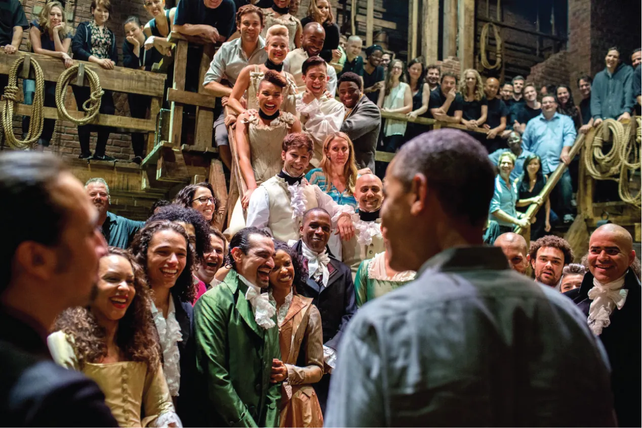 A photo of President Barack Obama meeting the cast of Hamilton. The cast members are in costume and are on the stage of the musical.