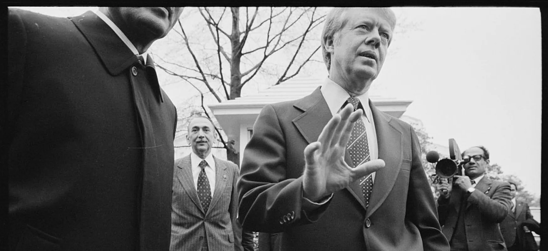 President Jimmy Carter stands outdoors, speaking, next to a man whose face is cut out of the photograph. A camera crew in the background films the president.